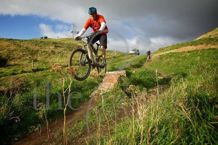 A mountain biker in an orange shirt and helmet is captured mid-air as he jumps off a small ramp, surrounded by lush green hills and a cloudy sky. A second cyclist is seen in the background, riding along a dirt path. Chimney Sweep mountain bike trail.