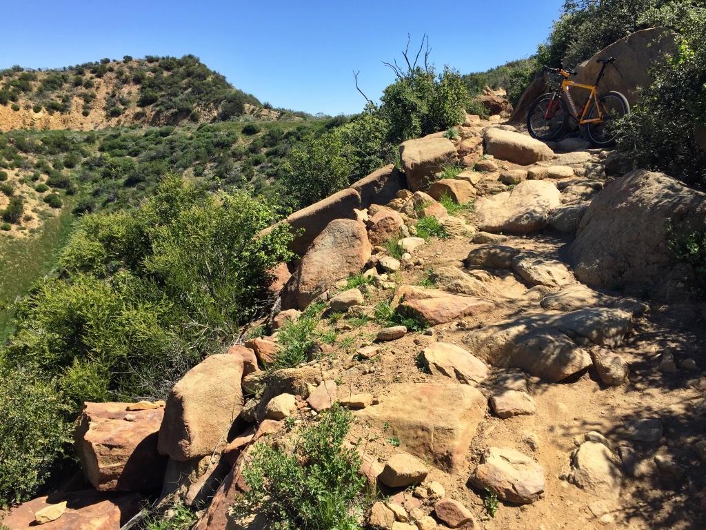 Mountain biking path with rocky terrain and greenery on the sides. An orange bicycle is parked nearby, surrounded by large stones and shrubs under a clear blue sky. Chumash mountain bike trail.