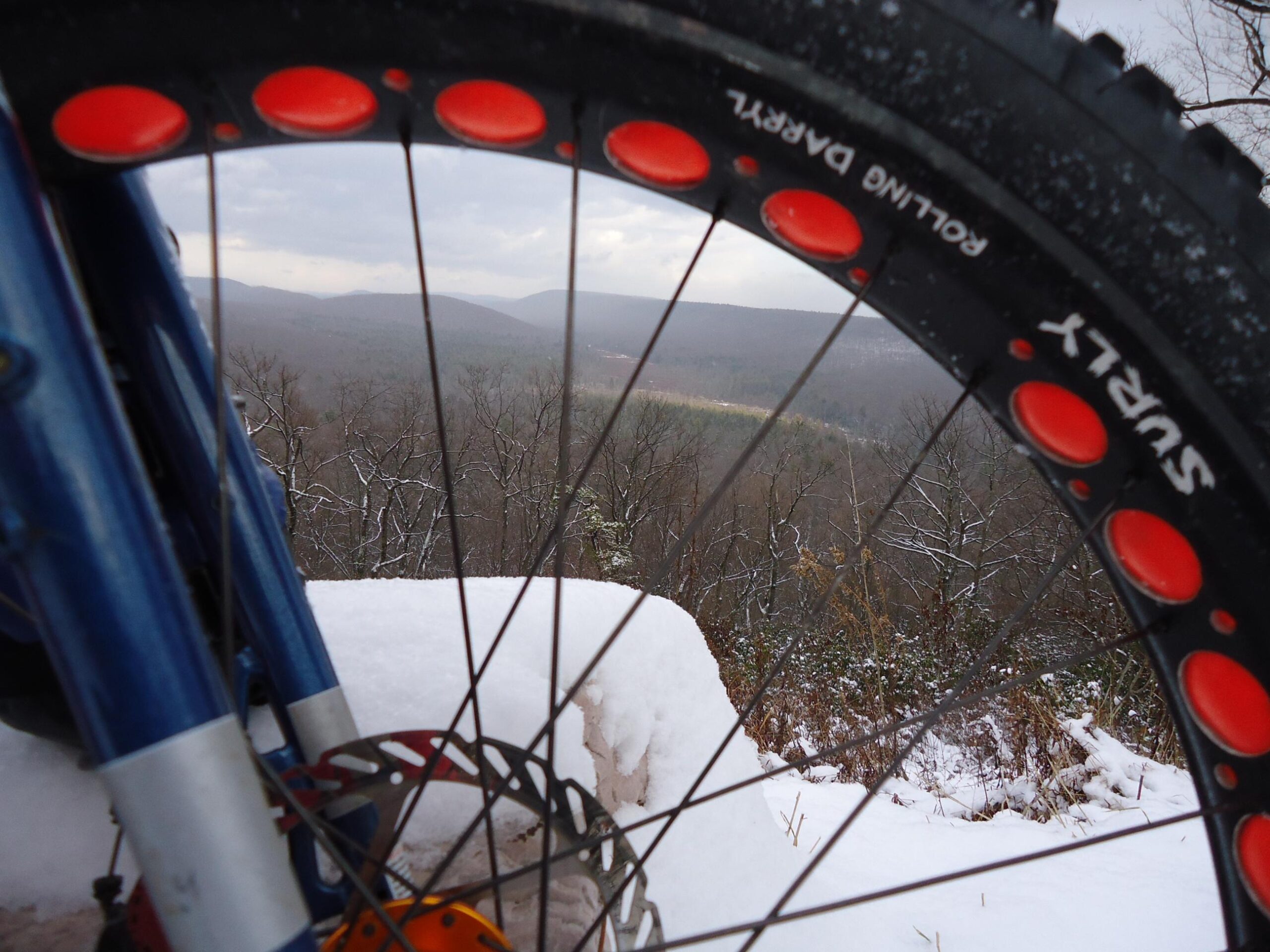 Salsa Mukluk 3: A close-up view of a bicycle wheel with a rugged tire, set against a snowy landscape. The background features a distant mountain range under a cloudy sky, while the foreground shows a dusting of snow on the ground. The bike's blue frame and orange components are partially visible alongside the wheel.