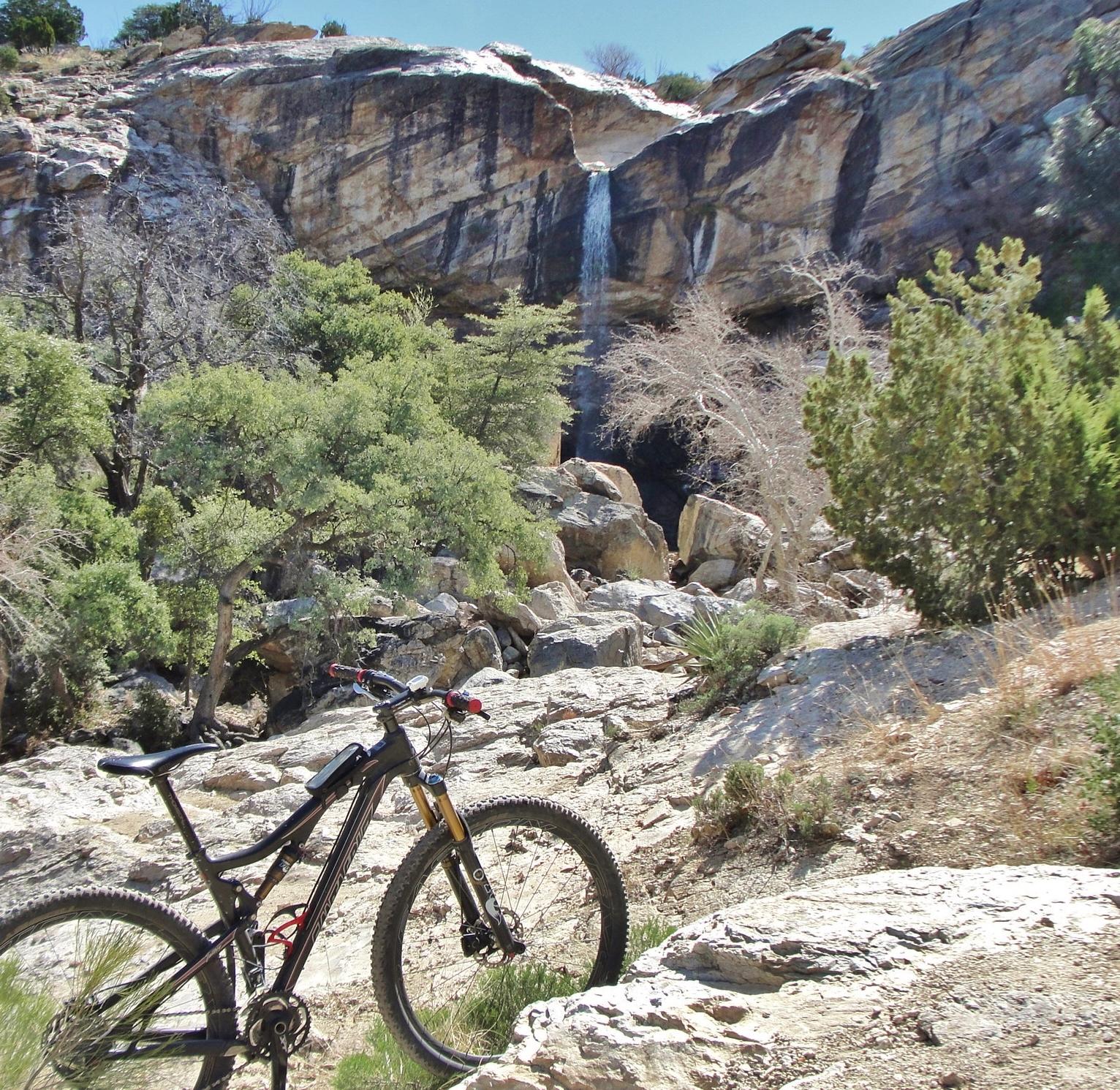 A mountain bike resting on rocky terrain in a natural landscape, featuring a waterfall and lush greenery in the background. Chiva Falls mountain bike trail.