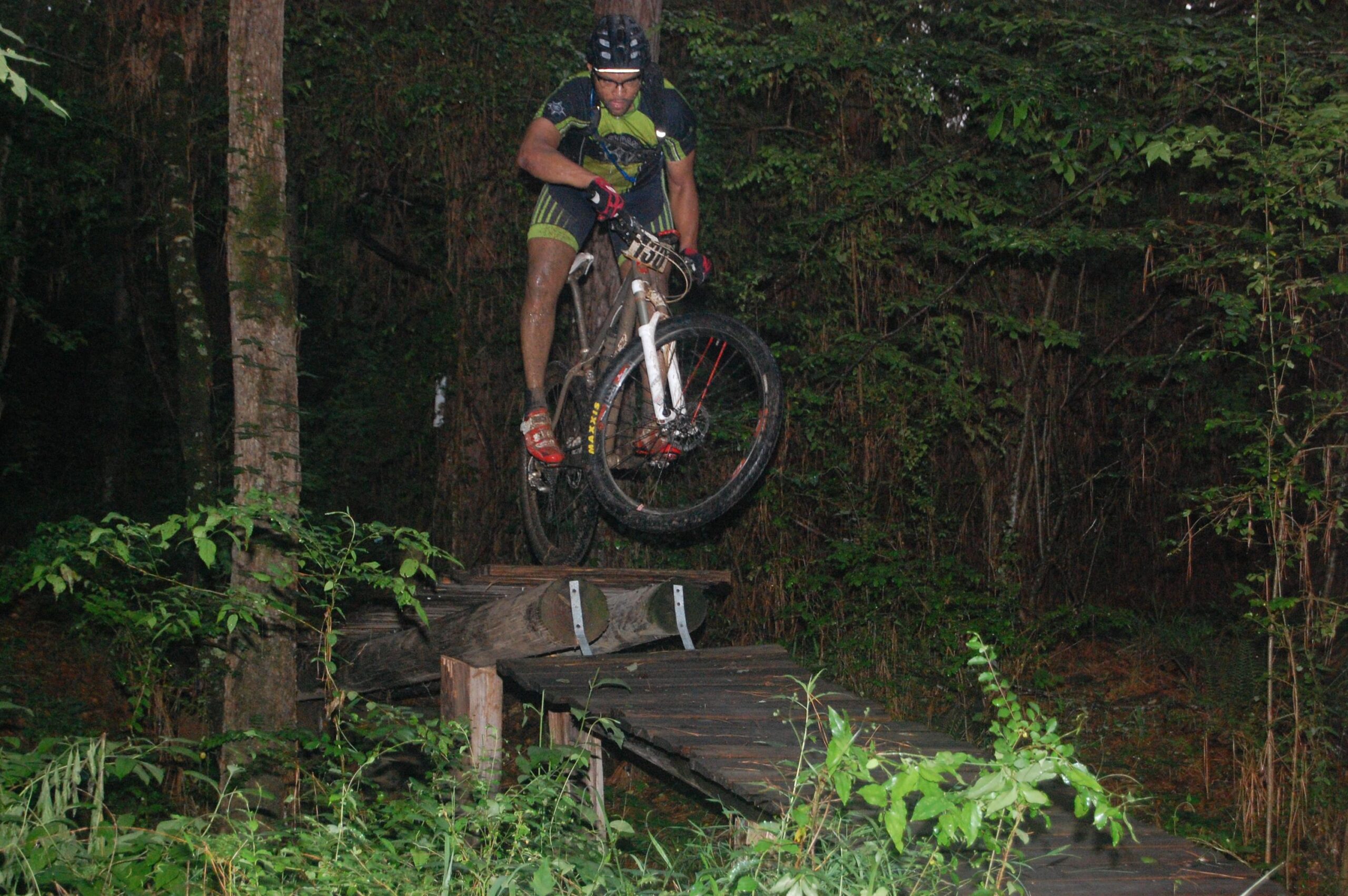 A mountain biker performing a jump off a wooden bridge in a dense forest. The cyclist is wearing a helmet and specialized biking gear, showcasing a dynamic action pose while airborne above the bridge. Surrounding vegetation includes trees and shrubs, contributing to the natural environment. Mt. Zion Bike Trails mountain bike trail.