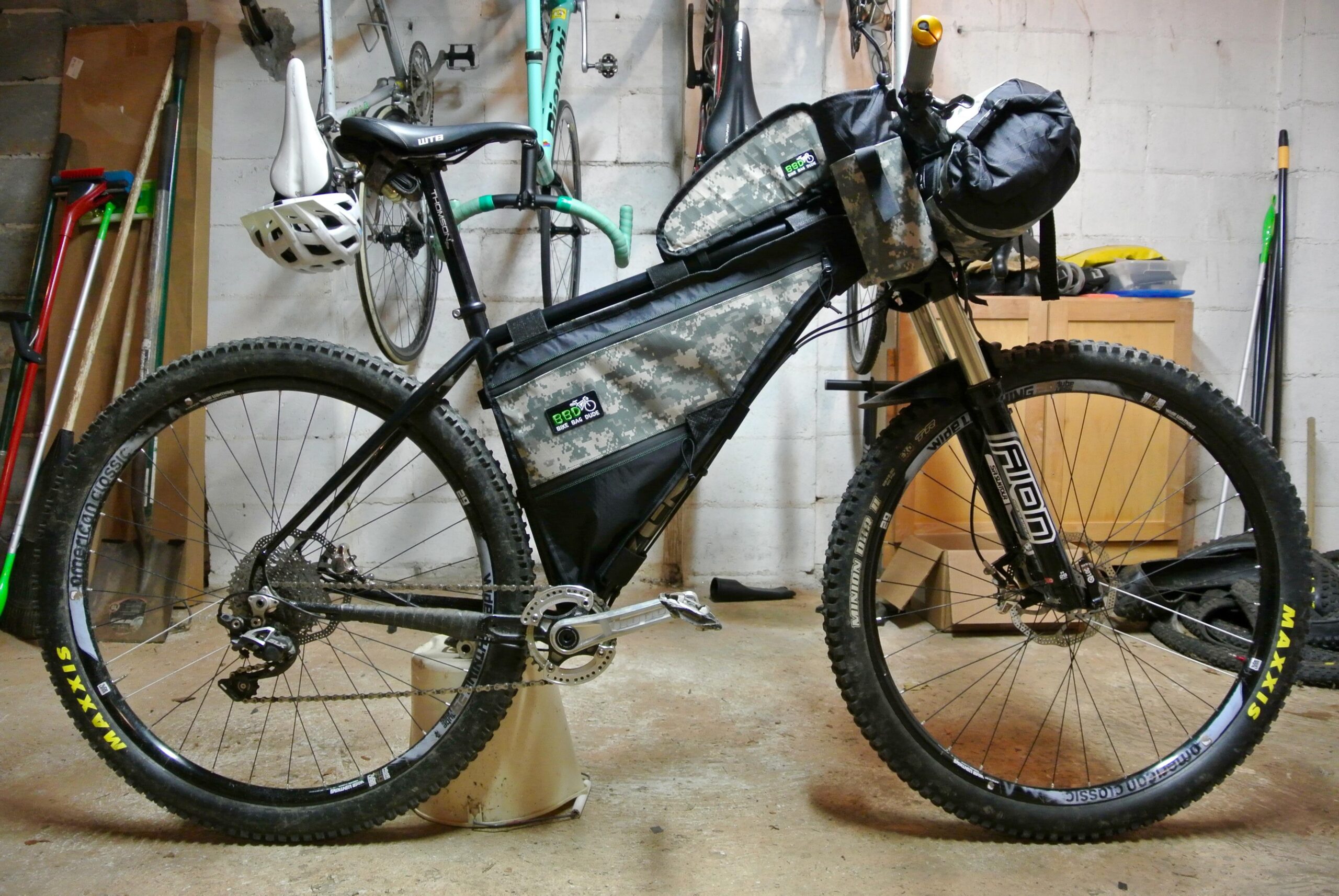Zen TRAIL Frame: A black mountain bike with a camouflage-patterned frame bag and a front carrying pouch, positioned in a garage. The background includes various tools and another bike hanging on the wall. The bike features thick tires and a sturdy frame, indicating a focus on off-road cycling.