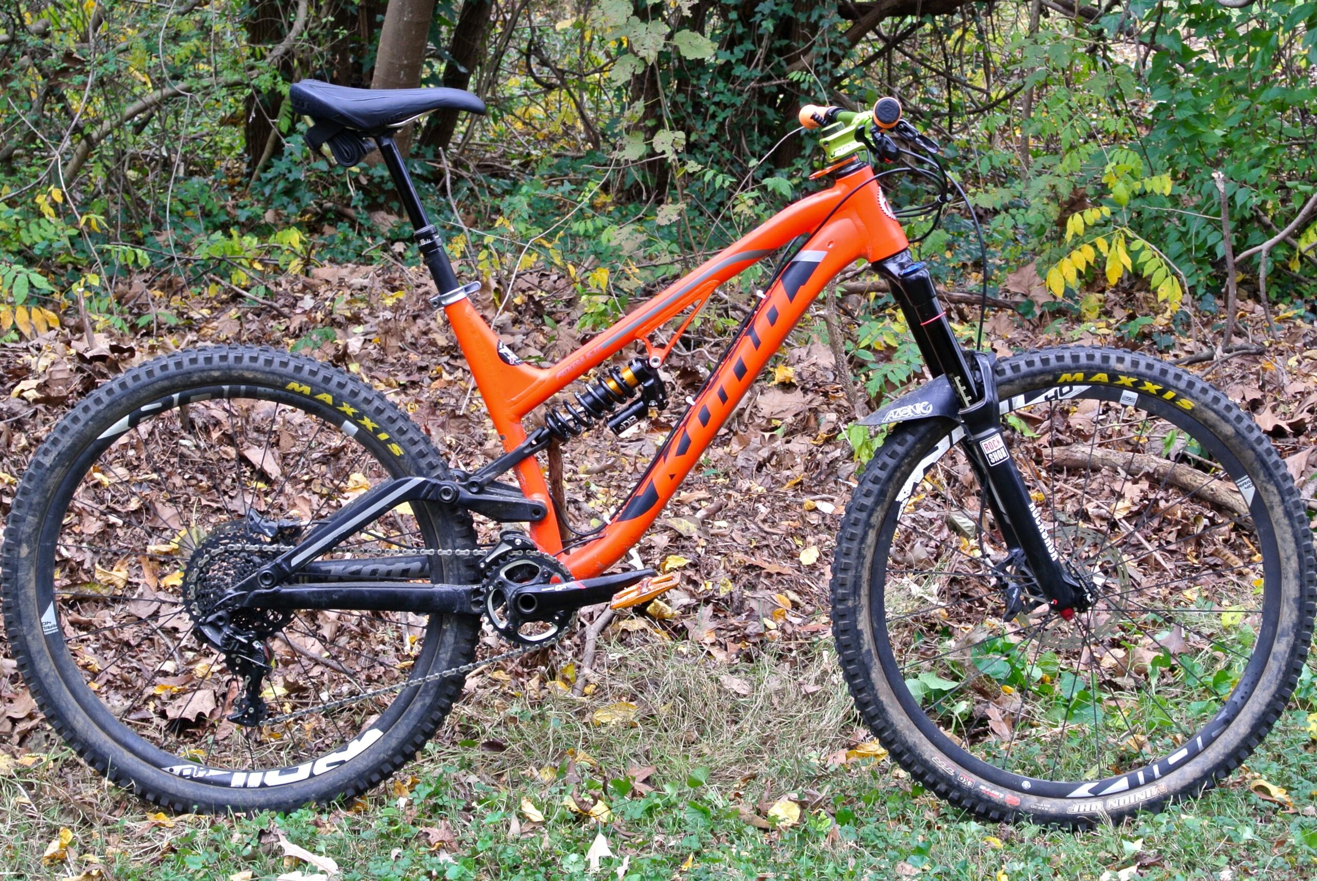 Kona Process 153 DL: A vibrant orange mountain bike with a sturdy frame, equipped with a rear shock, wide tires, and a black seat. It is positioned on a ground covered with fallen leaves, surrounded by greenery in the background.