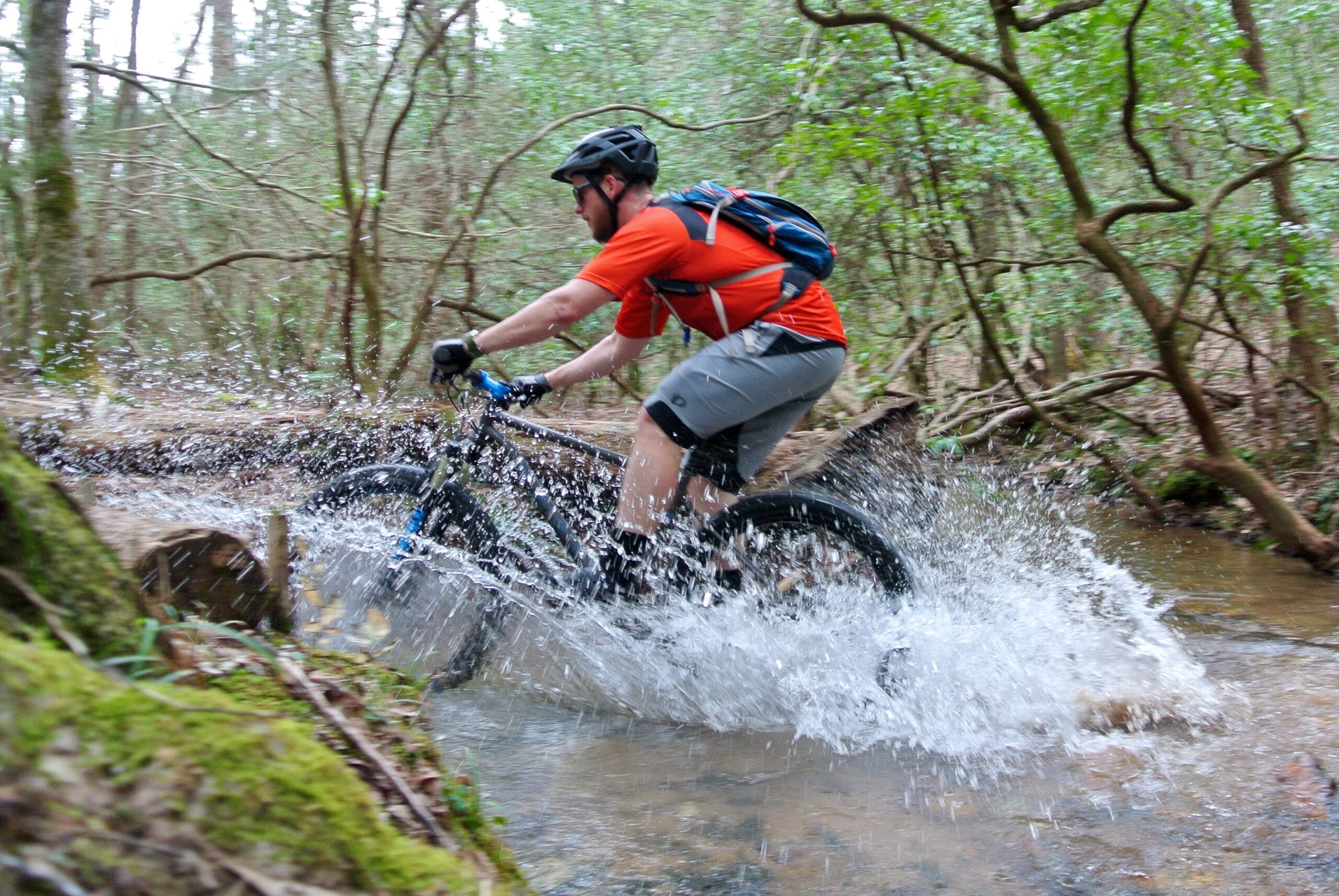 Marin Pine Mountain 2 27.5: A mountain biker in an orange shirt and gray shorts rides through a shallow stream, splashing water as they navigate the wooded trail surrounded by lush greenery.