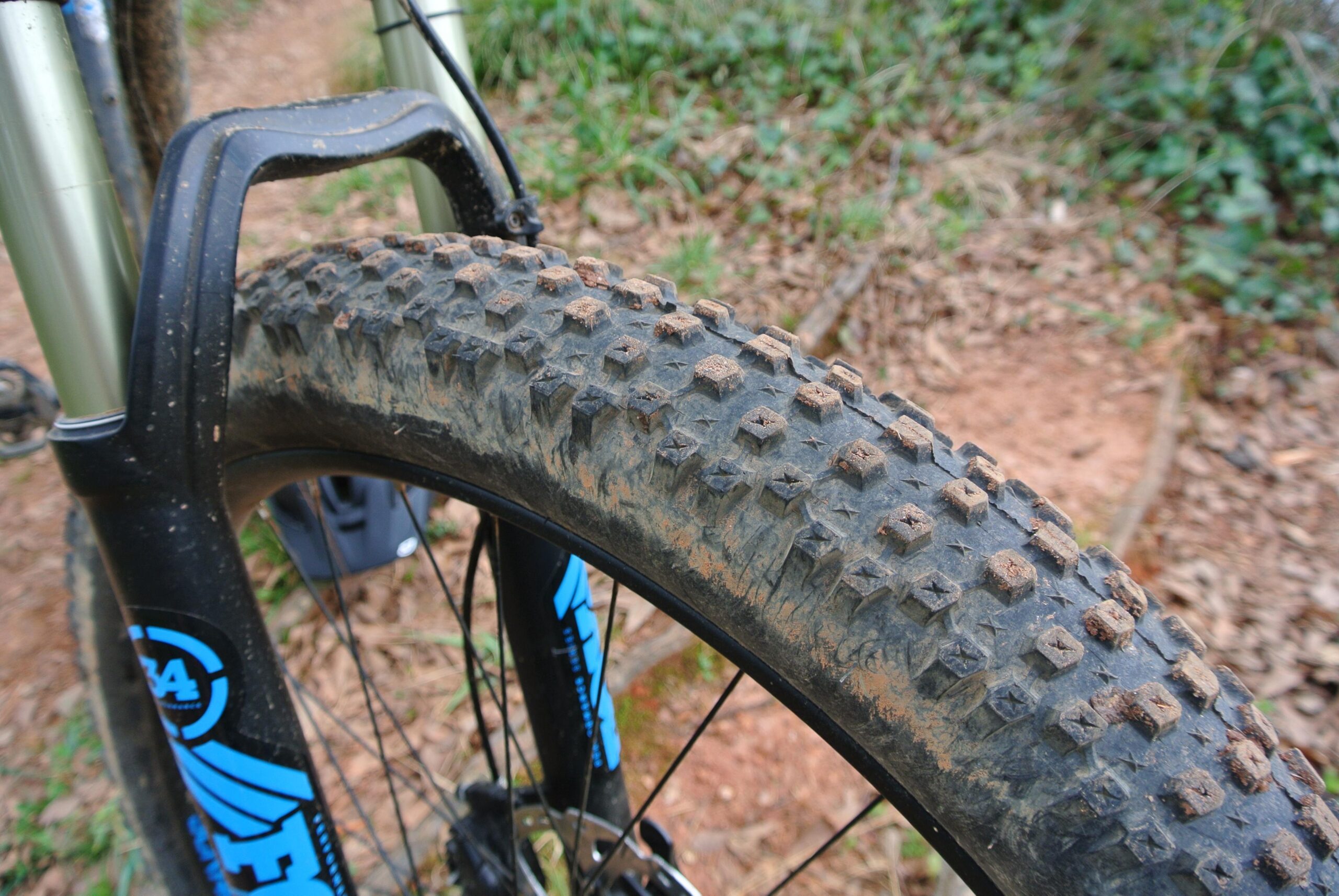 WTB Bridger: Close-up of a mountain bike tire showing its tread pattern, which is designed for traction on rough terrain. The tire is slightly muddy, indicating recent use on a trail, with rocky soil and foliage in the background.