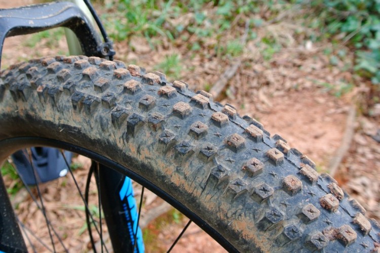 Close-up view of a mountain bike tire showing its textured surface, with visible dirt and mud particles, highlighting the tread pattern designed for traction on rugged terrain. The background features a blurred natural setting with foliage.