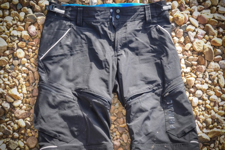 Black outdoor shorts laid out on a bed of pebbles, featuring a blue waistband and reflective details. The shorts have side pockets and appear designed for active use.