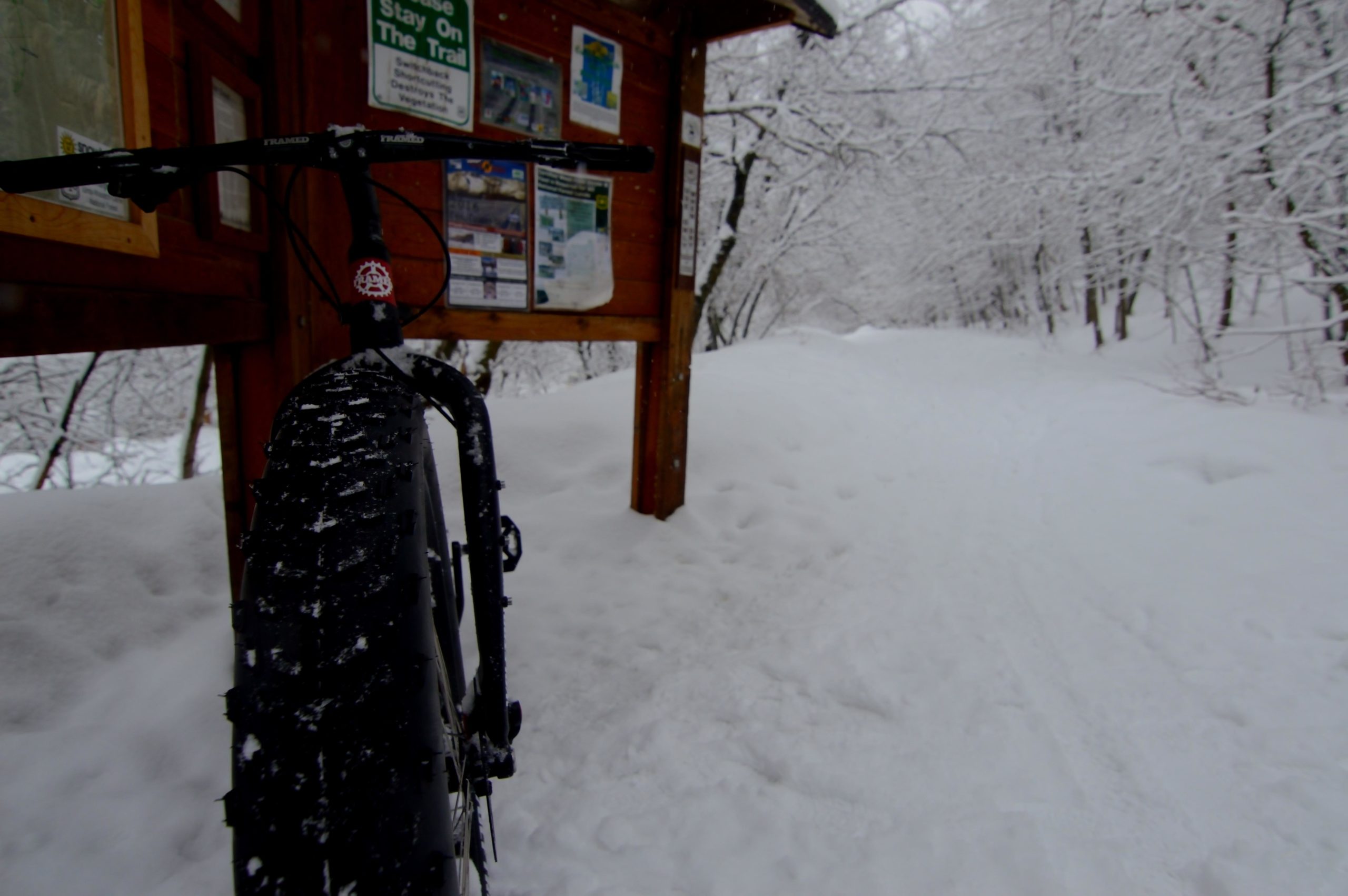 A fat tire bike parked next to a wooden trail sign, surrounded by a snowy landscape with trees covered in snow in the background. The trail is lightly marked in the snow. Bonneville Shoreline Trail - Ogden Section mountain bike trail.