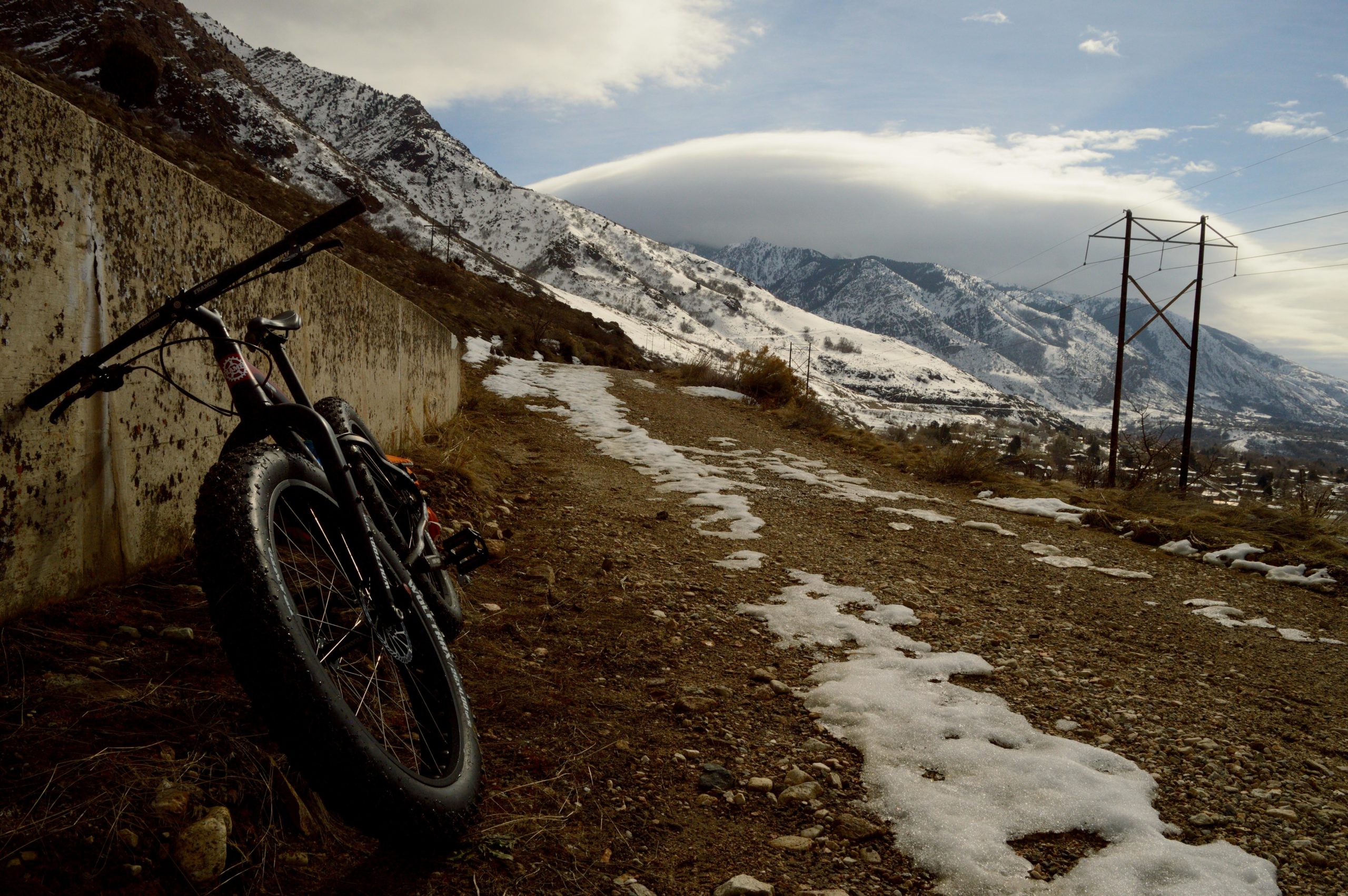 A fat-tire bike rests against a concrete wall on a gravel path, with snow patches visible along the trail. In the background, majestic snow-capped mountains stretch beneath a cloudy sky, while power lines run alongside the road. The scene captures a tranquil winter landscape. Bonneville Shoreline Trail - Ogden Section mountain bike trail.
