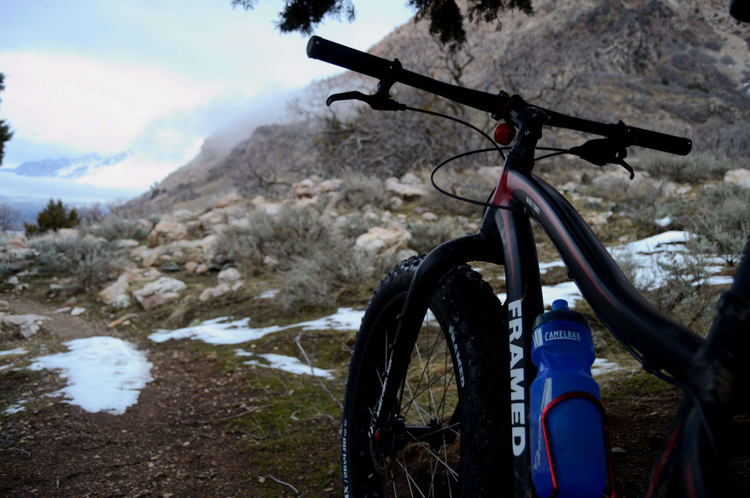 A fat tire mountain bike rests on a dirt and gravel path in a rugged outdoor setting. The bike is partially obscured in the foreground, showcasing its large tires and frame with the word "FRAMED." A blue water bottle is attached to the bike, and the background features rocky terrain with sparse vegetation and a cloudy sky, suggesting a mountainous area. Snow patches are visible on the ground, indicating a chilly environment. Bonneville Shoreline Trail - Ogden Section mountain bike trail.