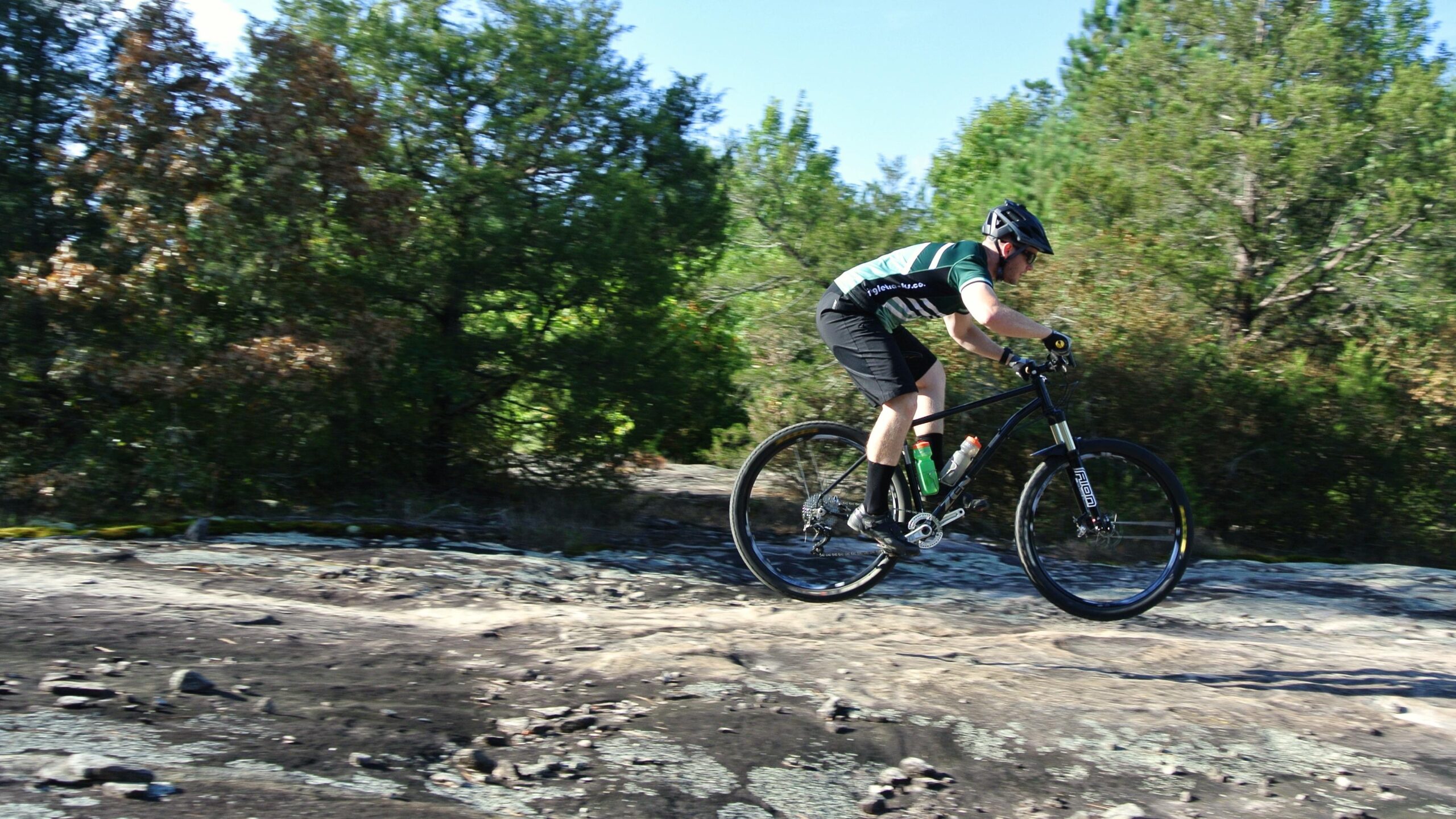 Zen TRAIL Frame: A cyclist in a black and green jersey performs a jump on a rocky outdoor trail, surrounded by trees and greenery on a sunny day.