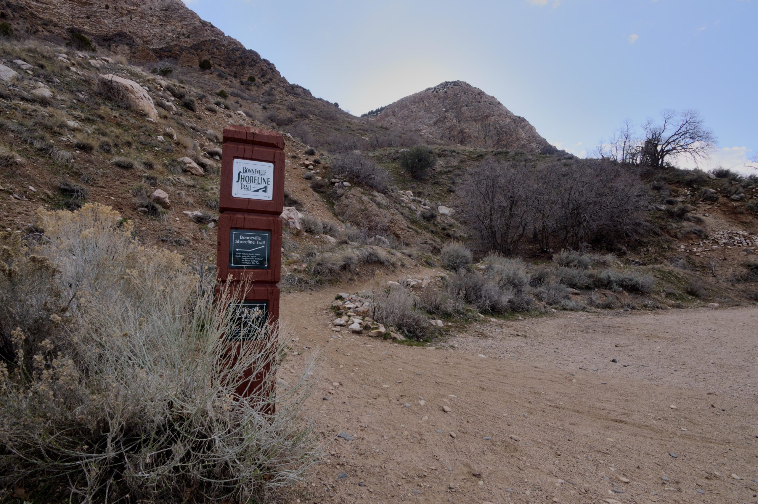 Sign for the Bonneville Shoreline Trail at the base of a rocky hillside, surrounded by sparse vegetation and gravel paths, under a cloudy sky. Bonneville Shoreline Trail - Ogden Section mountain bike trail.
