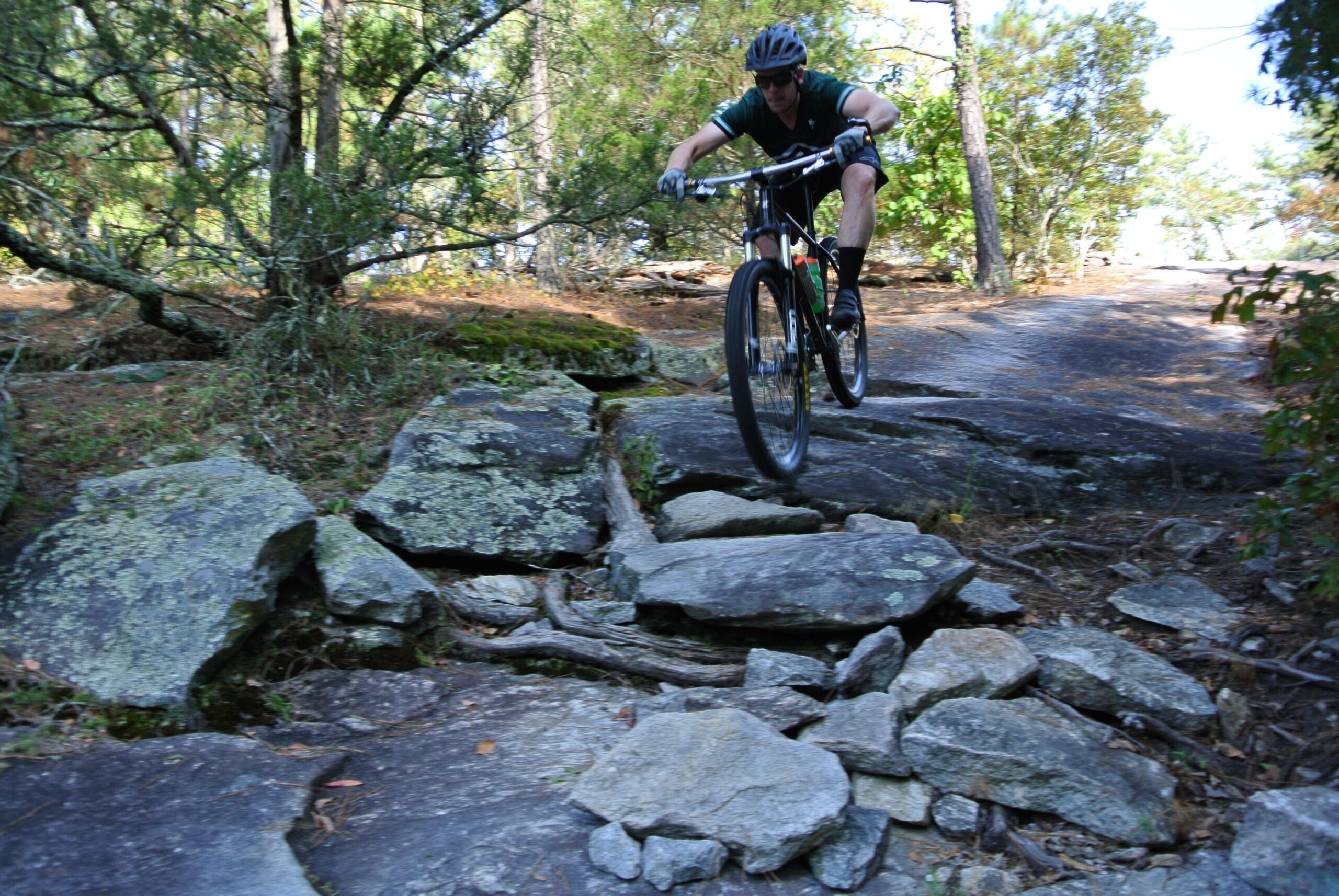 Zen TRAIL Frame: A mountain biker navigating a rocky trail, airborne as the bike lifts off a large rock. Lush green trees and foliage are present in the background, highlighting a natural outdoor setting. The rider is wearing a helmet and biking gear, focused on maintaining balance on the challenging terrain.