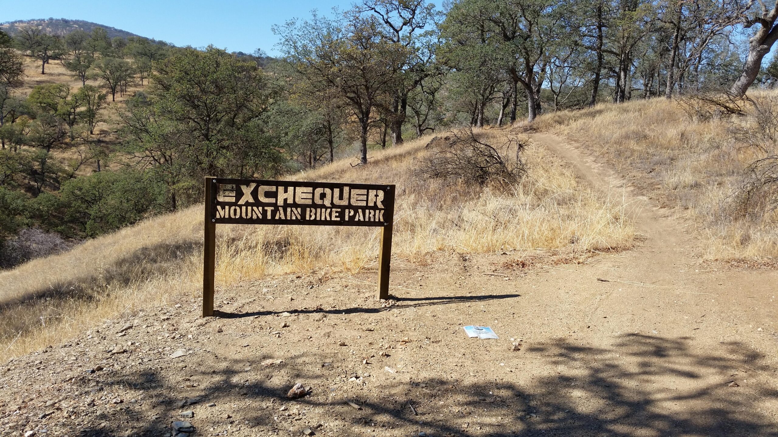 Sign for Exchequer Mountain Bike Park at the start of a dirt trail, surrounded by dry grass and trees on a sunny day. Exchequer mountain bike trail.