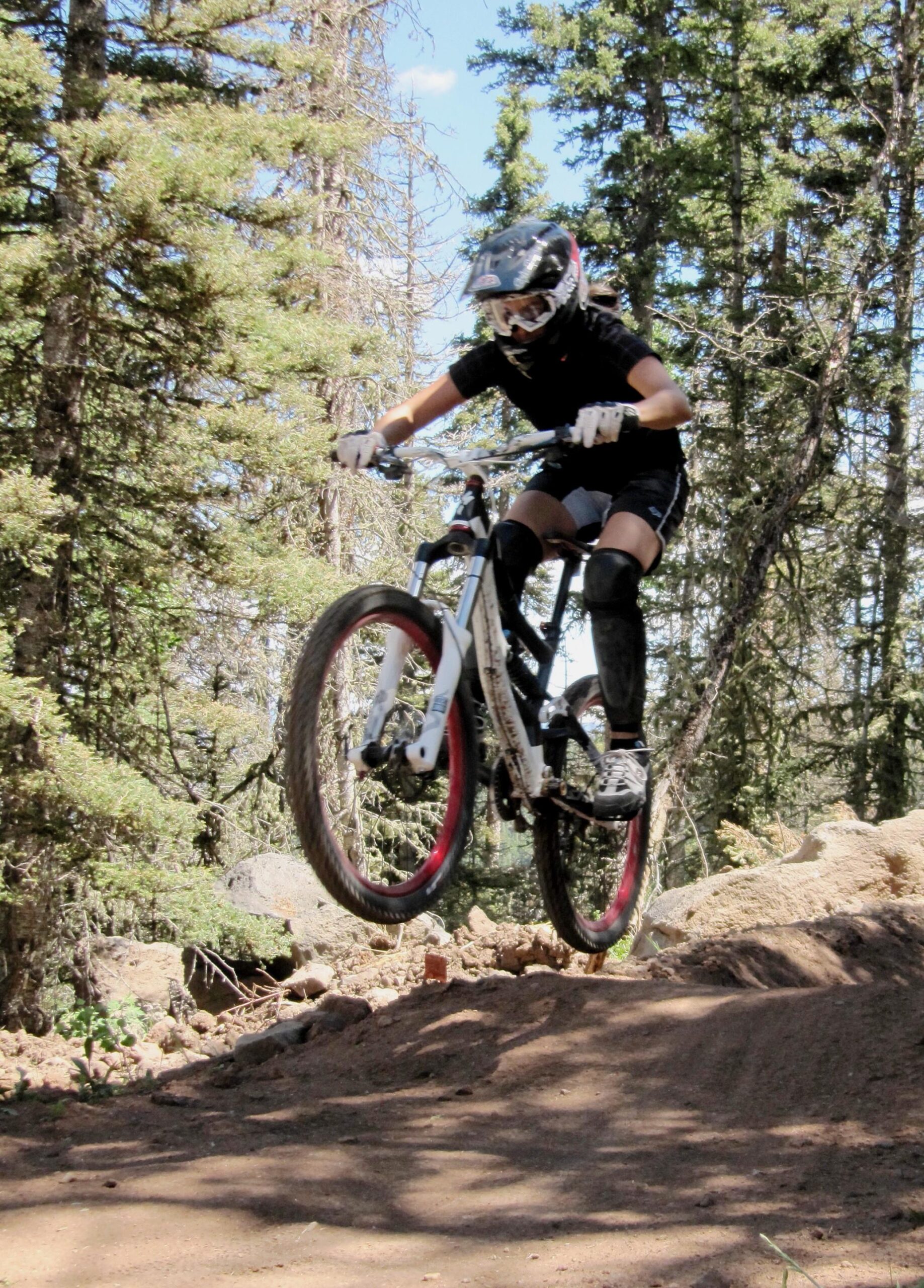 A person riding a mountain bike performs a jump on a dirt trail surrounded by trees, wearing a helmet and protective gear. The bike is airborne, with its front wheel elevated off the ground. Angel Fire Bike Park mountain bike trail.