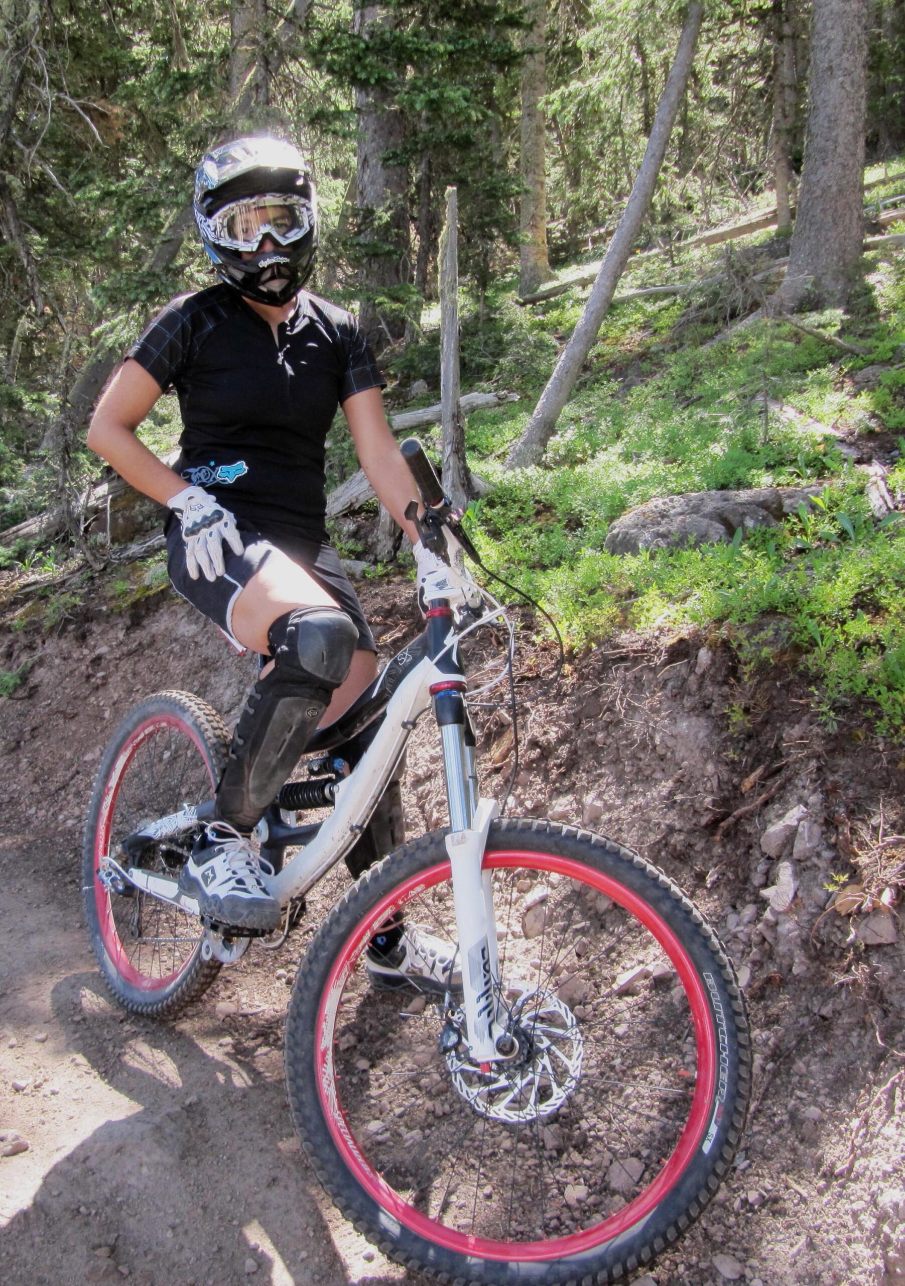 A person wearing a helmet, goggles, and protective gear is sitting on a mountain bike on a dirt trail surrounded by trees. The bike features white and red rims, and the rider is dressed in a short-sleeve black jersey and shorts, with knee pads visible. Sunlight filters through the trees, illuminating the scene. Angel Fire Bike Park mountain bike trail.