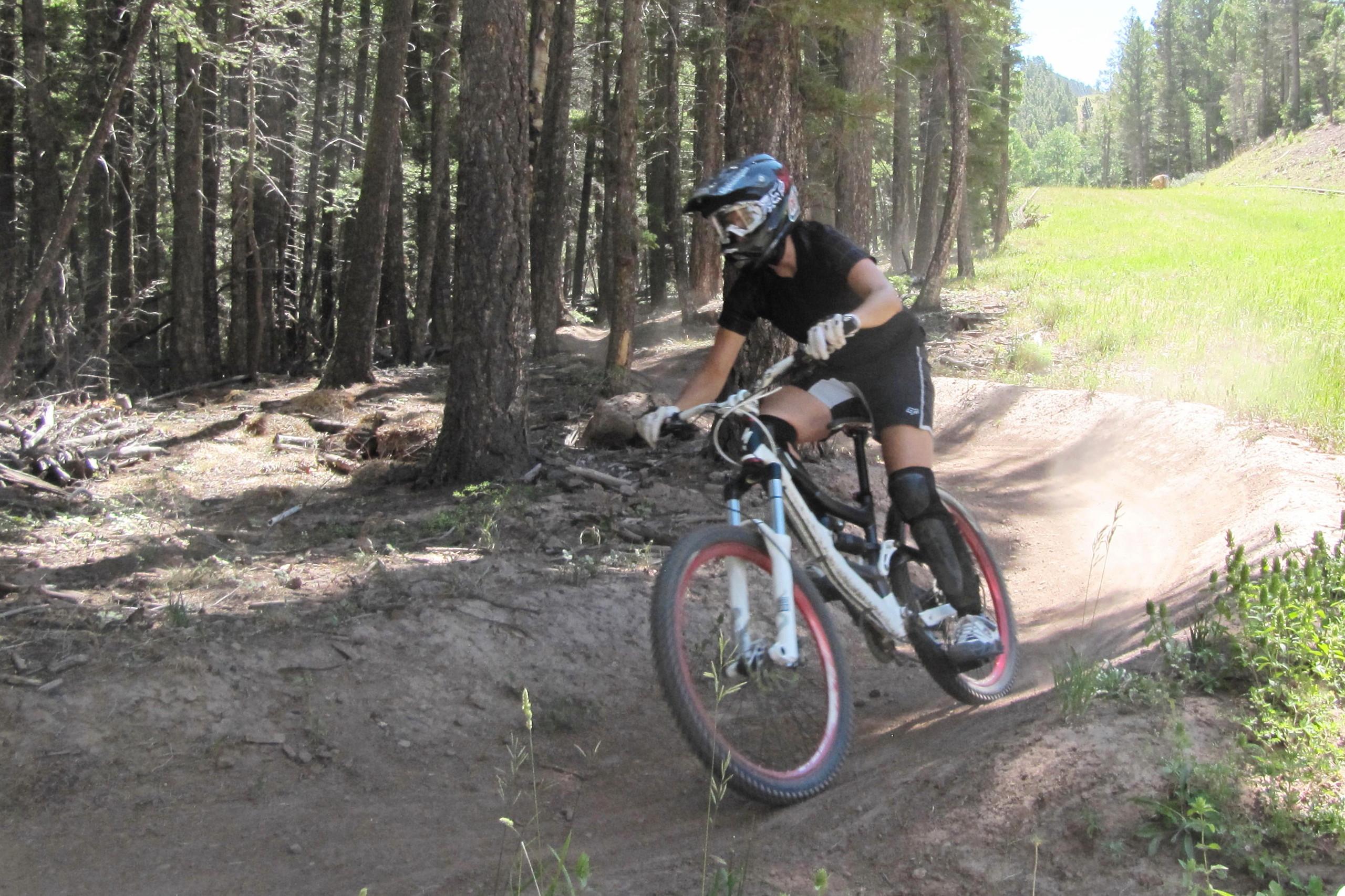 A mountain biker riding on a dirt trail through a forest, wearing protective gear and a helmet. Dust is kicked up from the trail as they navigate a curve surrounded by tall trees and green grass. Angel Fire Bike Park mountain bike trail.