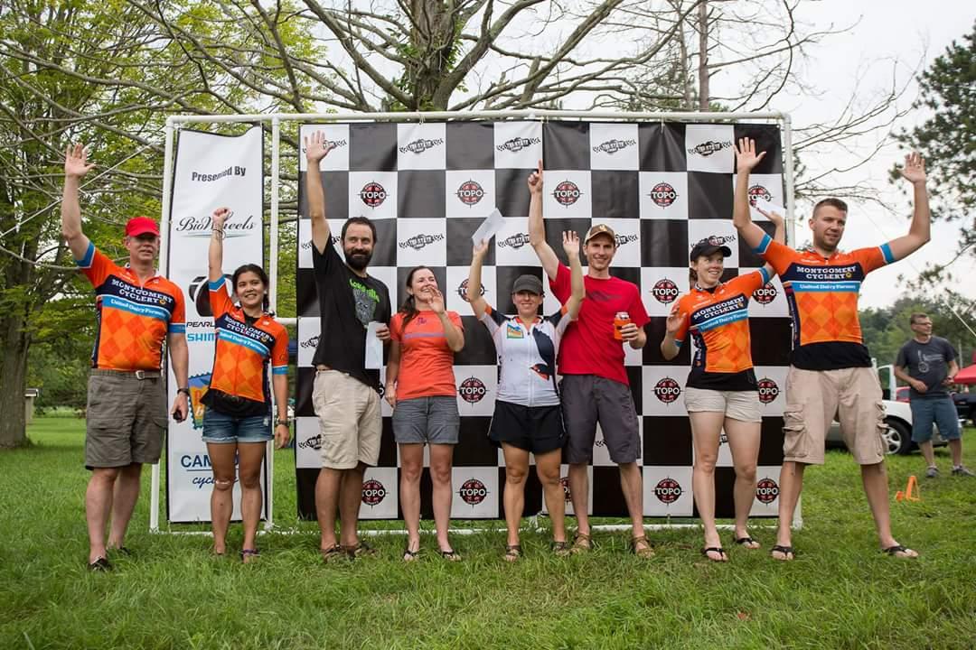 A group of eight cyclists stands on a podium, smiling and waving to the camera. They are dressed in colorful cycling jerseys, with a mix of shorts and casual attire. The backdrop features a black and white checkered design with sponsor logos, and they appear to be celebrating a cycling event or competition. The setting is outdoors, with trees in the background and a sunny atmosphere. Hueston Woods State Park mountain bike trail.