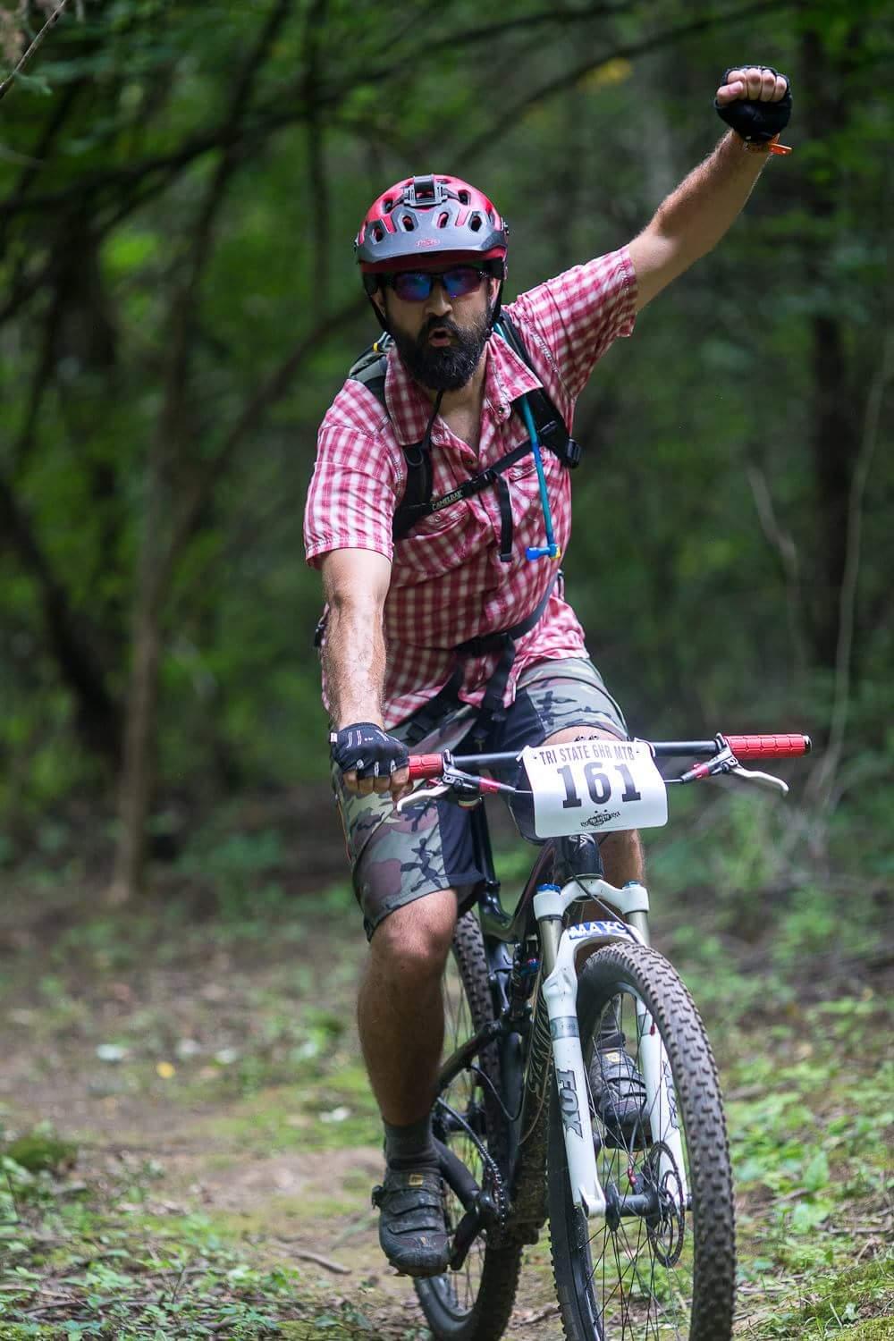 A mountain biker wearing a red helmet and sunglasses raises one arm in celebration while riding on a trail surrounded by greenery. He is dressed in a plaid shirt and camouflage shorts, with a race number pinned to his bike. The scene captures a moment of excitement and achievement in a natural setting. Hueston Woods State Park mountain bike trail.