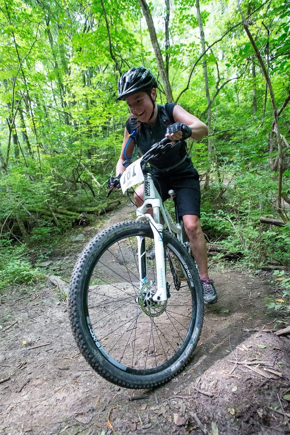 A person mountain biking on a narrow trail in a lush green forest. The cyclist is wearing a helmet and a black outfit, appearing focused as they navigate over a rocky section of the path. The terrain is natural and surrounded by trees, showcasing a vibrant and adventurous outdoor environment. The bike has a white frame with a visible race number attached. Hueston Woods State Park mountain bike trail.