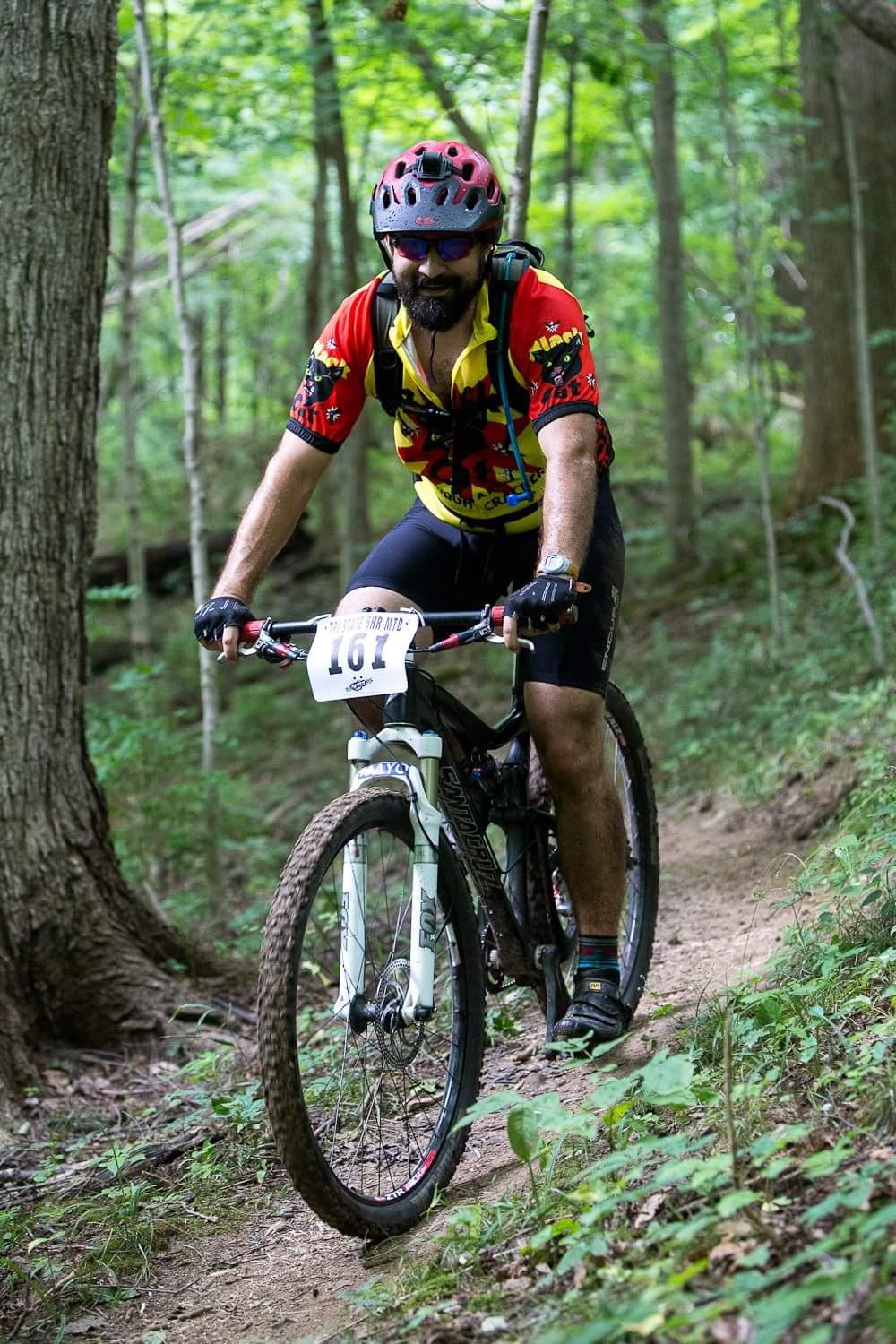 A person riding a mountain bike on a dirt trail surrounded by green trees, wearing a red and yellow cycling jersey, black shorts, and a helmet. The cyclist has a beard and is focused on navigating the trail, with a race number attached to the bike. Hueston Woods State Park mountain bike trail.