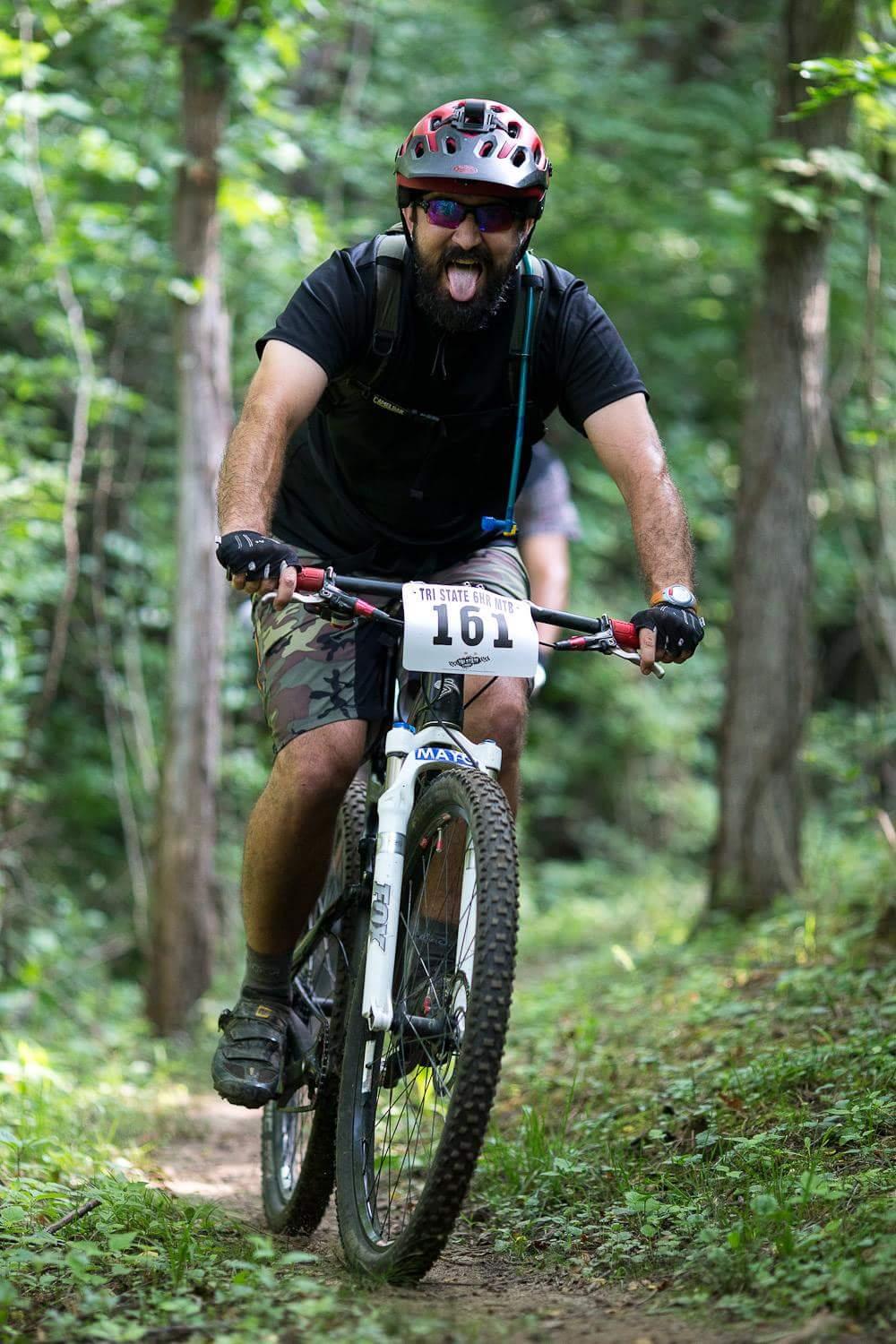 A mountain biker sticks out his tongue while riding on a dirt trail surrounded by lush green trees. He is wearing a black helmet and sunglasses, along with a black shirt and camouflage shorts. A race number is displayed on his bike. Hueston Woods State Park mountain bike trail.