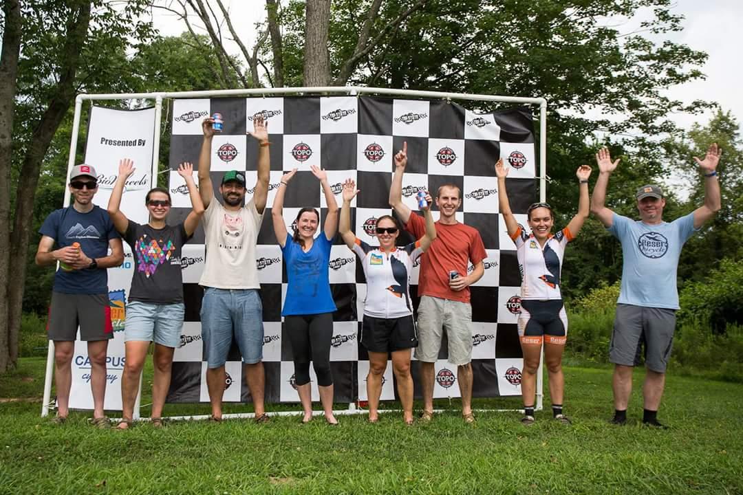 A group of eight people standing together outdoors in front of a checkered backdrop, celebrating with their arms raised. Various individuals are wearing casual and athletic clothing, some holding beverages. The setting appears to be a festive outdoor event, with trees and grass in the background. England Idlewild Mountain Biking Park mountain bike trail.