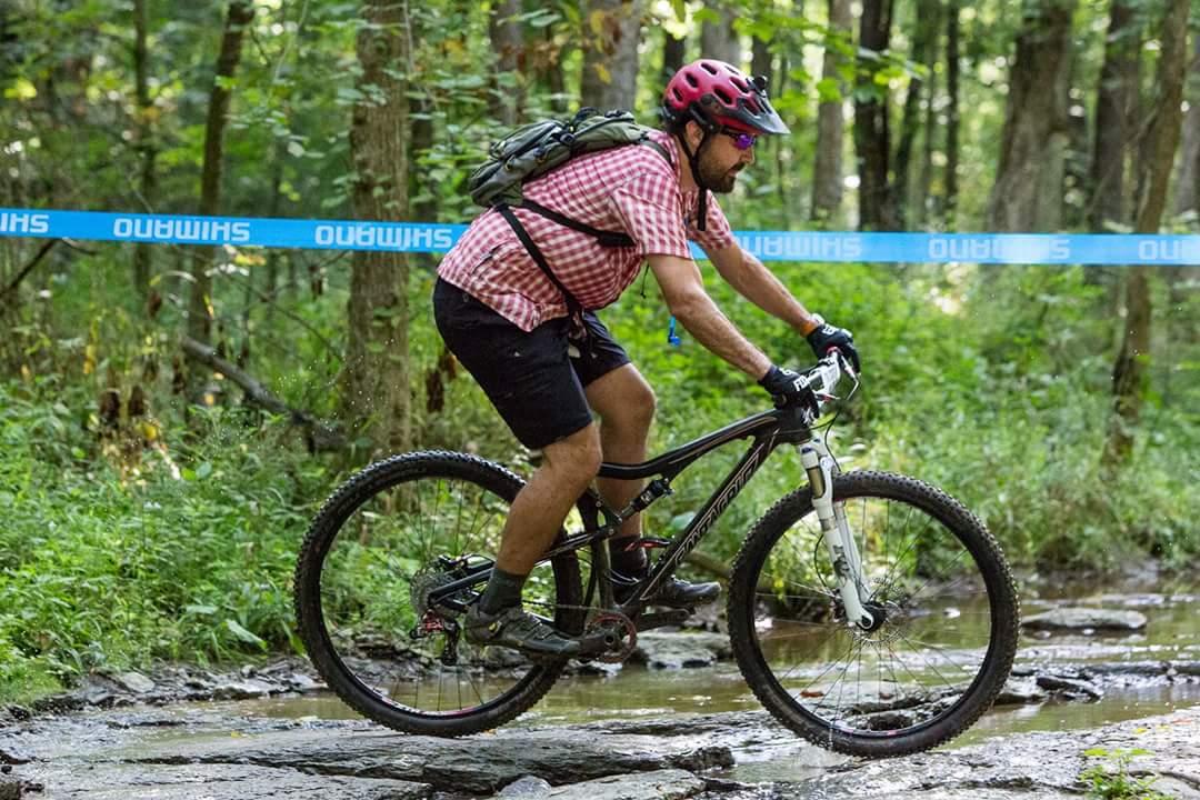 A mountain biker wearing a patterned shirt and a helmet rides through a rocky area with splashes of water, surrounded by green trees. A blue banner in the background displays the Shimano logo. England Idlewild Mountain Biking Park mountain bike trail.