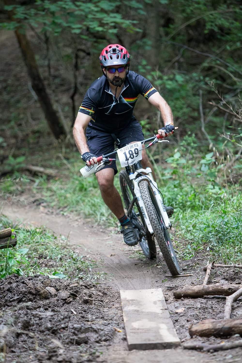 A mountain biker navigates a dirt trail in a forest, riding over a wooden plank as part of a race. He wears a black cycling jersey with colorful stripes, a helmet, and sunglasses. The trail is surrounded by greenery and muddy sections. The rider’s race number, 189, is visible on his bike. England Idlewild Mountain Biking Park mountain bike trail.