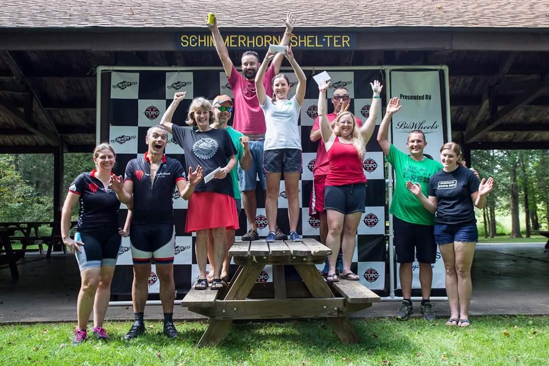 A group of people celebrating together on a picnic table, with some holding trophies or awards, in front of a checkered backdrop that reads “Presented By [Sponsor Name].” The setting appears to be outdoors under a shelter, with trees visible in the background, and the participants are smiling and waving, showcasing a joyful atmosphere. Versailles State Park mountain bike trail.