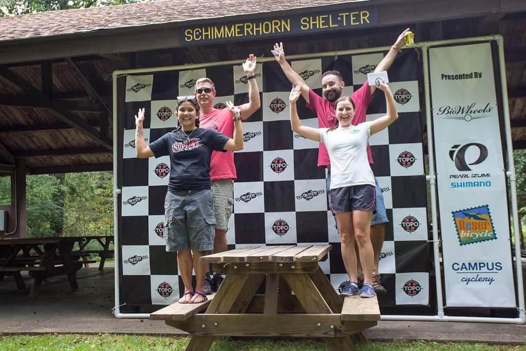 Four participants celebrate on a wooden platform after an event, raising their arms in joy. The backdrop features a black and white checkered banner with the text "SCHIMMERHORN SHELTER" and various sponsor logos. The setting appears to be outdoors, likely at a park or recreational area. Versailles State Park mountain bike trail.