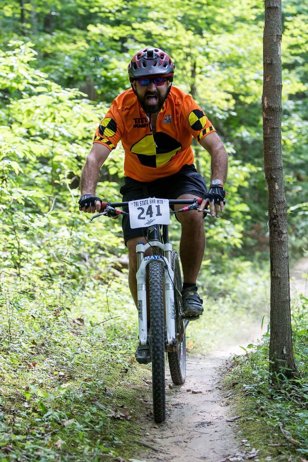 A man riding a mountain bike on a dirt trail through a forest, wearing an orange jersey with a black design and helmet. He is showing an intense expression, possibly shouting or cheering, as he navigates the path. The surrounding greenery includes trees and plants, indicating a vibrant, natural environment. A race number is visible on his jersey. Versailles State Park mountain bike trail.