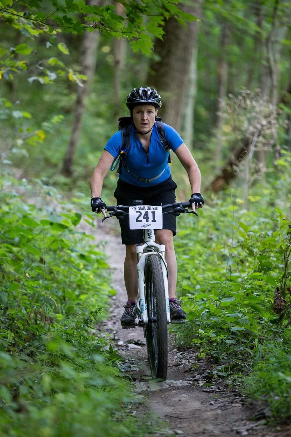 A woman riding a mountain bike on a narrow dirt trail surrounded by greenery, wearing a blue shirt and a helmet. She has a race number pinned to her bike and appears focused as she navigates the trail. Versailles State Park mountain bike trail.