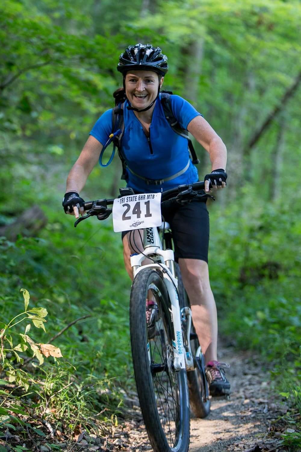 A smiling woman in a blue cycling jersey rides a mountain bike on a dirt trail surrounded by greenery. She is wearing a helmet and gloves, and her bike has a race number pinned to it. Versailles State Park mountain bike trail.
