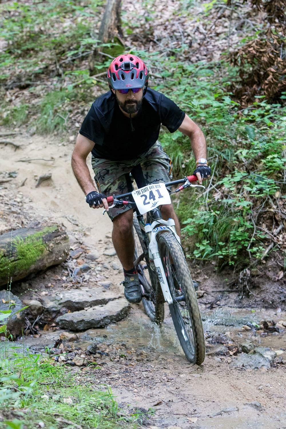 A mountain biker wearing a red and black helmet navigates a rocky trail surrounded by lush greenery. The biker is focused, leaning forward on a white mountain bike as they ride over a stream of water on the path. The cyclist is wearing a black t-shirt and camouflage shorts, showcasing an active outdoor scene. Versailles State Park mountain bike trail.