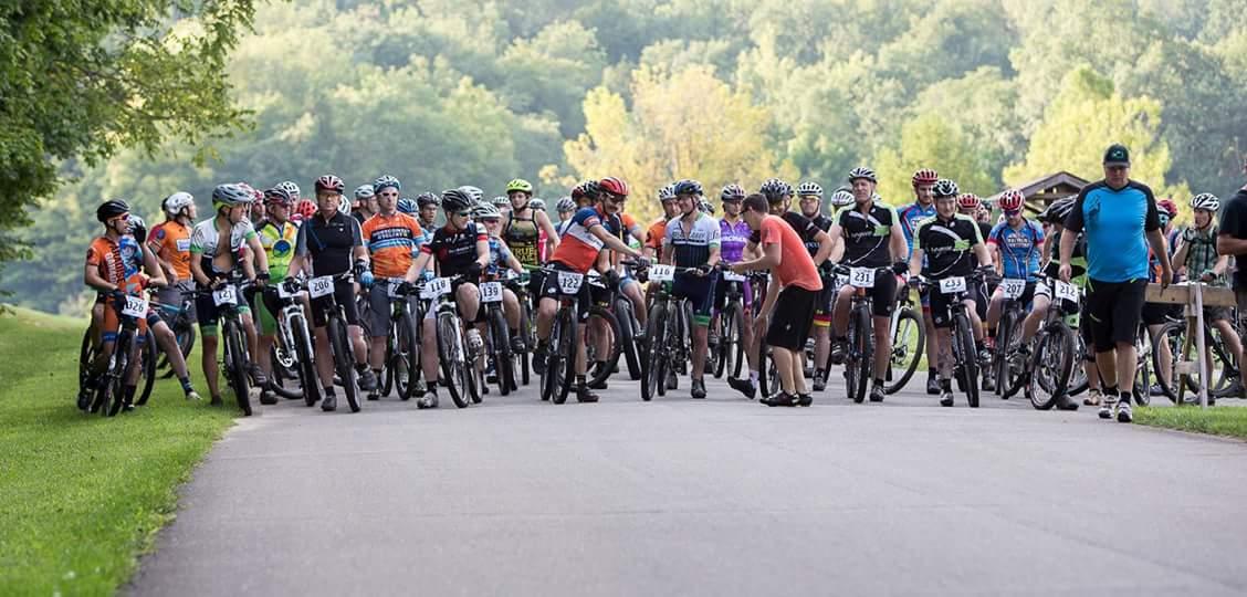 A group of cyclists wearing helmets and colorful jerseys gather on a paved road, preparing for a race or event. The scene is set in a natural outdoor environment with greenery in the background, suggesting a lively and competitive atmosphere. Versailles State Park mountain bike trail.