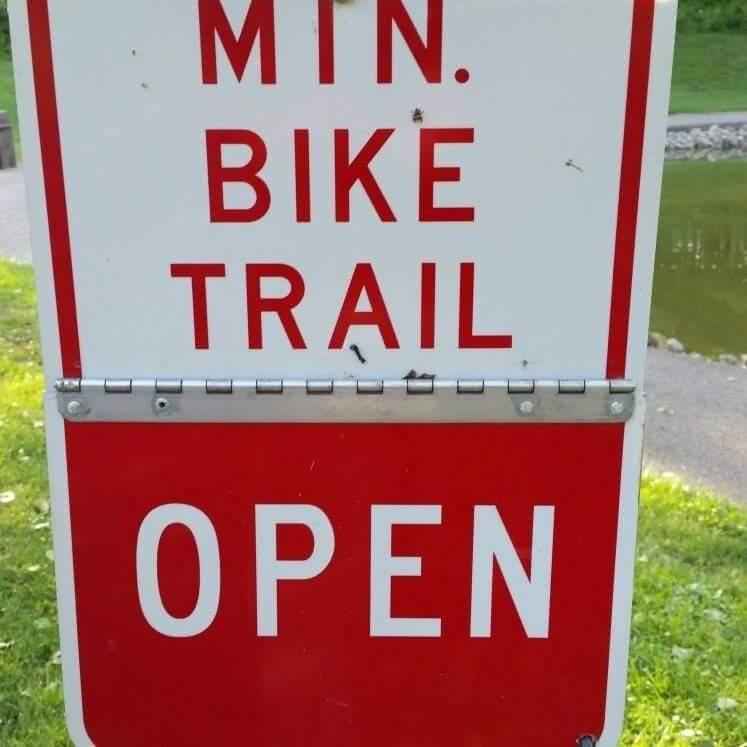 A sign indicating that a bike trail is open, featuring bold red and white letters that read "MIN. BIKE TRAIL OPEN." The sign is mounted on a grassy area with a water body visible in the background. Mitchell Memorial Forest Mountain Bike Trail mountain bike trail.