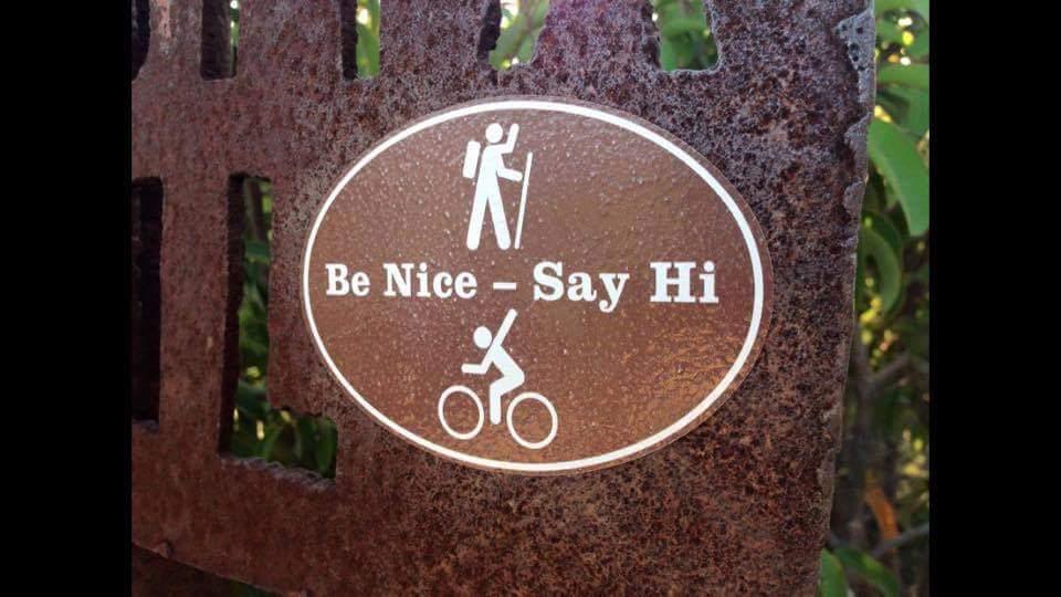 A metal sign featuring an illustration of a hiker and a cyclist, with the text "Be Nice - Say Hi" in between them. The sign is set against a natural background, suggesting a trail or recreational area. Mitchell Memorial Forest Mountain Bike Trail mountain bike trail.