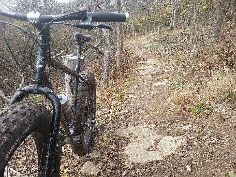 A close-up view of a black mountain bike parked on a rocky dirt trail, surrounded by autumn foliage and trees. The bike features wide tires and a water bottle mounted on the frame. The winding trail ahead is lined with scattered rocks and patches of grass. Mitchell Memorial Forest Mountain Bike Trail mountain bike trail.