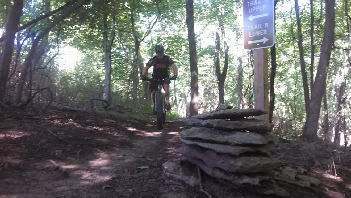 A mountain biker navigating a dirt trail in a wooded area, with a trail sign indicating the directions for "Trail B Upper" and "Trail B Lower" nearby. A stack of flat stones is positioned along the trail. The scene is bright, with sunlight filtering through the trees. Mitchell Memorial Forest Mountain Bike Trail mountain bike trail.