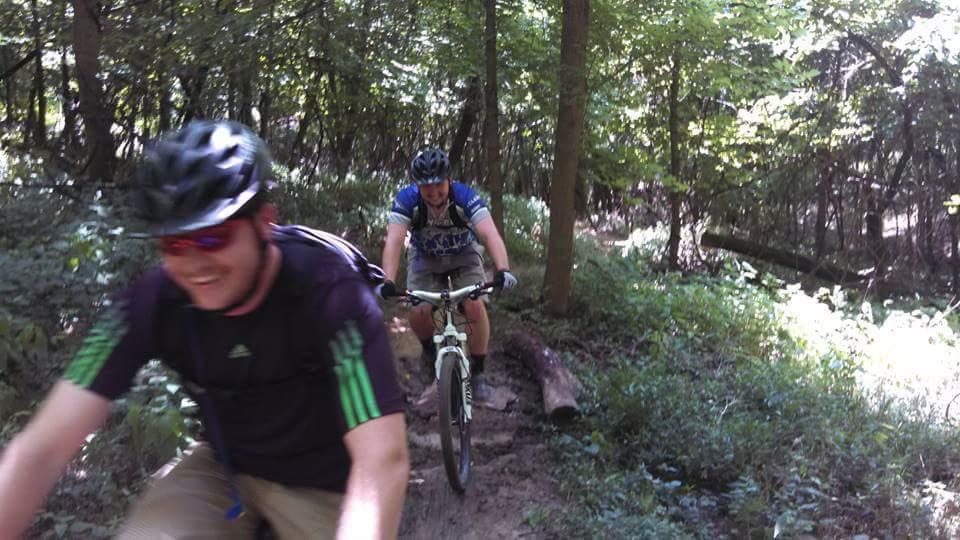 Two mountain bikers riding on a dirt trail through a lush green forest, with sunlight filtering through the trees. The person in the foreground is smiling and wearing a black shirt and helmet, while the second rider, slightly behind, is dressed in a blue and white jersey. Both are navigating the trail surrounded by foliage and natural scenery. Mitchell Memorial Forest Mountain Bike Trail mountain bike trail.