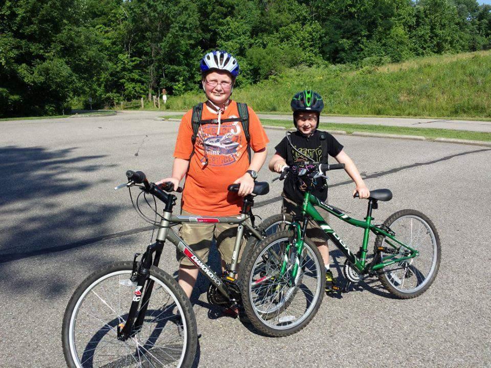 Two boys are standing with their bicycles on a sunny day in a park setting. One boy is wearing an orange T-shirt and a helmet, holding onto a silver bicycle, while the other boy, dressed in a black shirt and a green helmet, is next to a green bike. They both appear to be smiling and enjoying their time outdoors. Trees and grassy areas can be seen in the background. Mitchell Memorial Forest Mountain Bike Trail mountain bike trail.