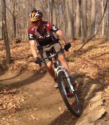 A mountain biker maneuvering on a dirt trail through a wooded area, surrounded by autumn leaves. The rider is wearing a helmet and cycling gear, leaning into a curve on the bike. Mitchell Memorial Forest Mountain Bike Trail mountain bike trail.