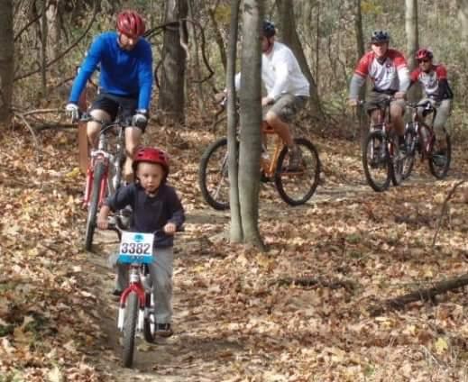 A young child in a red helmet rides a small bicycle on a dirt trail surrounded by autumn foliage, while several adults on mountain bikes ride in the background. The scene captures a family-friendly outdoor biking event. Mitchell Memorial Forest Mountain Bike Trail mountain bike trail.