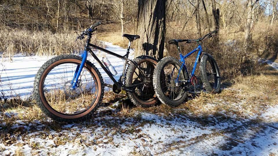 Two mountain bikes leaning against a tree along a snowy path, surrounded by bare trees and sparse vegetation. One bike is black with thick tires and a water bottle attached, while the other is blue with wide, knobby tires suitable for rough terrain. The ground is partially covered in snow, indicating a winter setting. Mitchell Memorial Forest Mountain Bike Trail mountain bike trail.