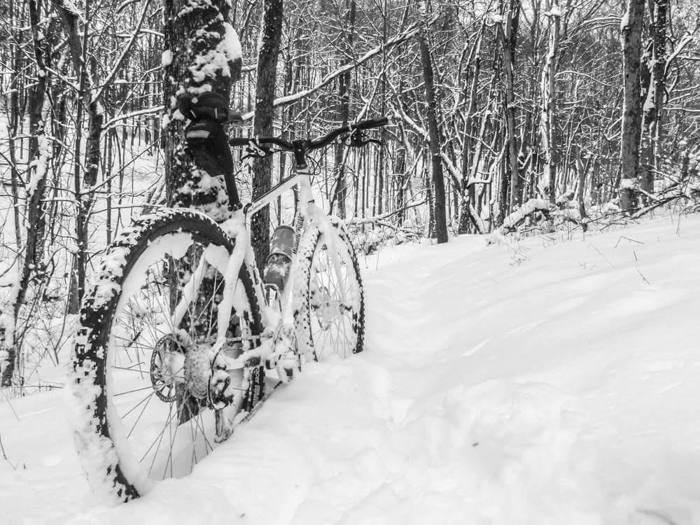 A mountain bike partially covered in snow, leaning against a tree in a snowy forest setting. The scene is in black and white, showcasing a quiet, winter landscape with trees blanketed in snow. The path is visible leading into the woods, emphasizing the wintry conditions. Mitchell Memorial Forest Mountain Bike Trail mountain bike trail.