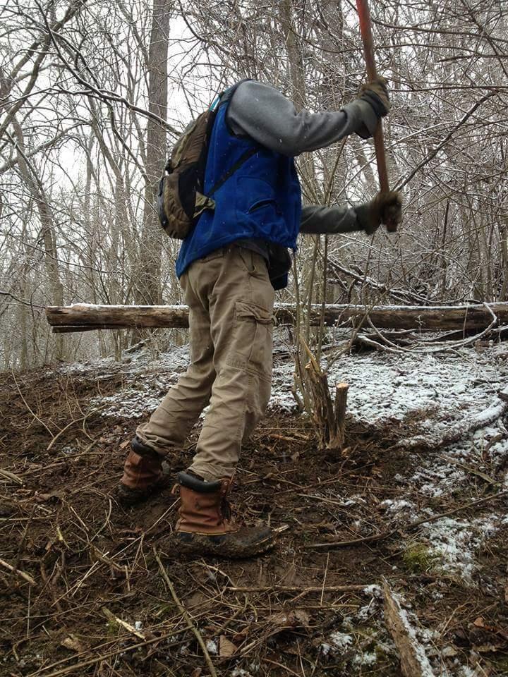 A person in outdoor work attire, including a blue vest and brown boots, is using a tool in a snowy, wooded area. The ground is mostly bare with some remnants of snow, and trees are visible in the background. The individual appears to be engaged in activities related to land management or outdoor maintenance. Hueston Woods State Park mountain bike trail.
