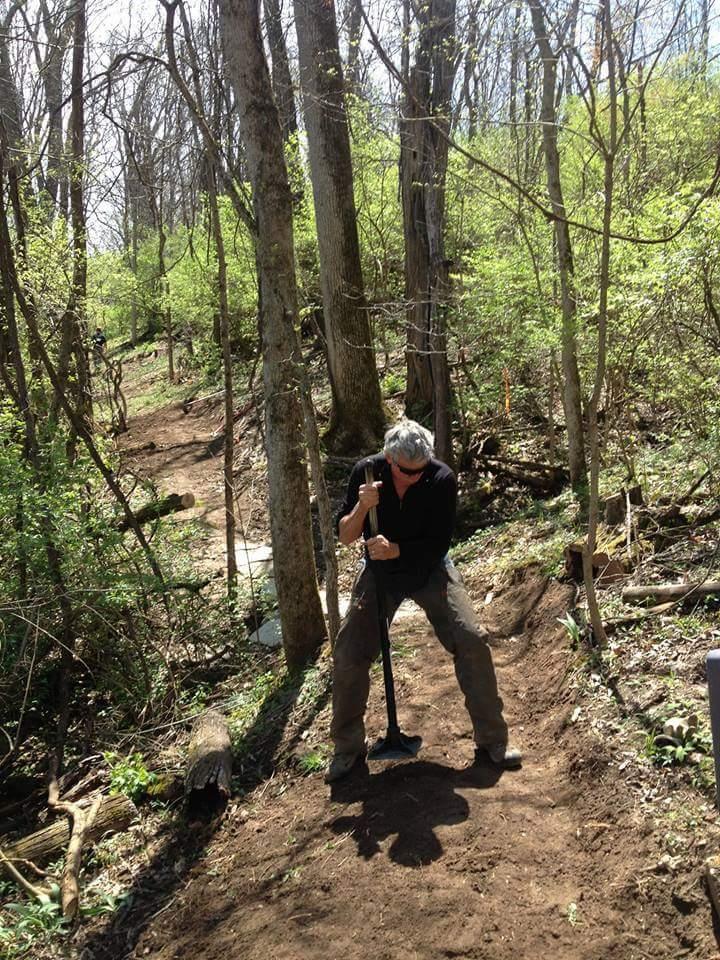 A person using a digging tool to work on a path in a wooded area, surrounded by trees and greenery. Sunlight filters through the branches, indicating a clear day. Hueston Woods State Park mountain bike trail.
