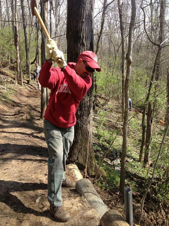 A person wearing a red hoodie and a cap, holding a large tool, is preparing to swing it while standing on a dirt path in a wooded area. Trees are visible in the background, and another individual can be seen further down the path. The scene suggests outdoor work or trail maintenance. Hueston Woods State Park mountain bike trail.