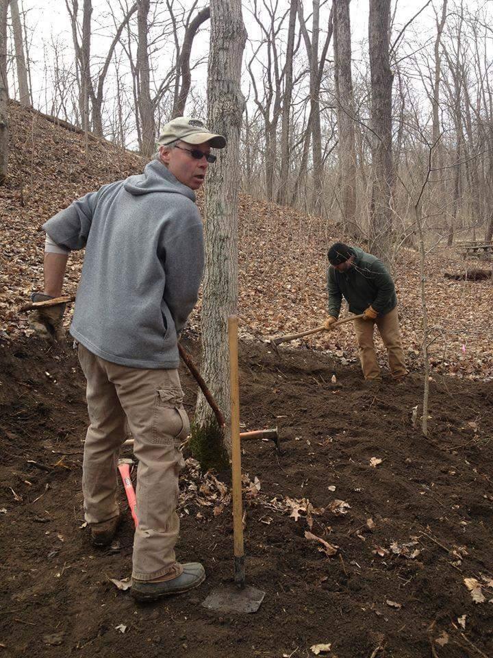 Two individuals working outdoors in a wooded area, one looking towards the camera while holding a digging tool, and another member focused on digging with a hoe. The scene is set in early spring with leafless trees and dry leaves covering the ground. Hueston Woods State Park mountain bike trail.